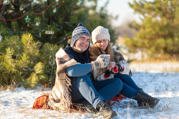 Jovem casal apaixonado bebe uma bebida quente, sentado no inverno na floresta, dobrado quente