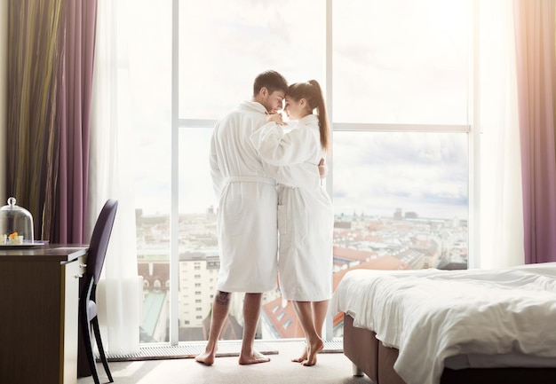 Jovem casal apaixonado em um quarto de hotel pela manhã, em pé na janela e se abraçando