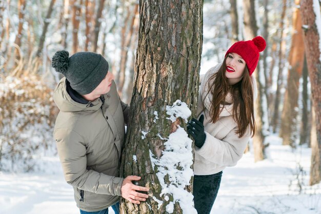 Jovem casal apaixonado espiando por trás de uma árvore na floresta de neve Sunny winter park