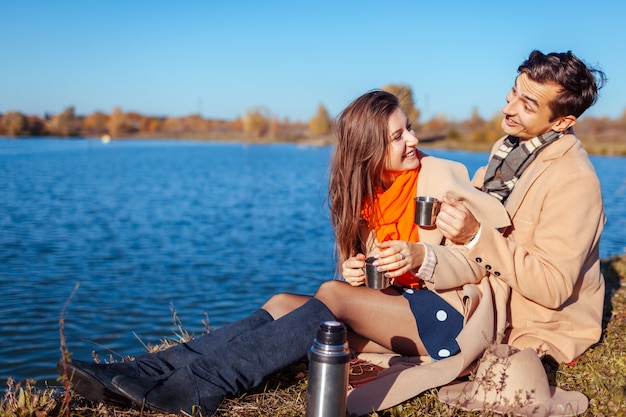 Jovem casal apaixonado fazendo piquenique no lago de outono. feliz homem e mulher brincando e curtindo chá