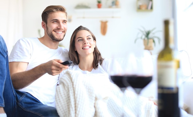 Jovem casal apaixonado no sofá em casa assistindo tv e rindo, bebendo uma taça de vinho tinto.