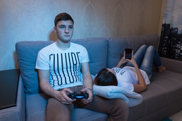 Jovem casal apaixonado passa o tempo junto enquanto o jovem joga videogame em casa com a esposa, que está deitada no sofá e navega na Internet pelo celular em frente a uma TV em casa