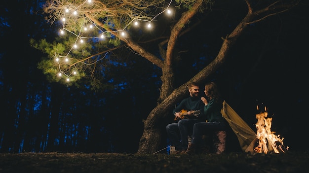 Jovem casal apaixonado por acampar turistas sentados perto de uma fogueira contra uma barraca na floresta com uma guirlanda retrô, foto com muito barulho, foco seletivo