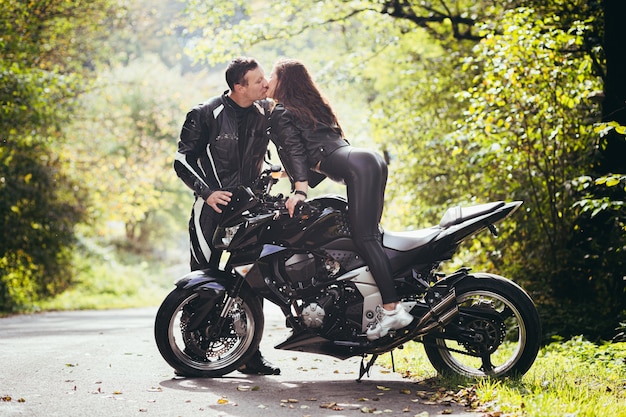 Foto jovem casal apaixonado se abraçando e beijando perto de uma motocicleta esportiva preta no fundo de uma estrada na floresta