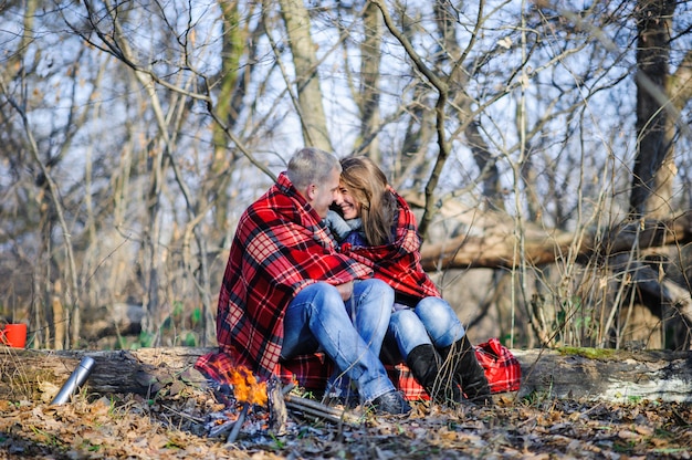 Jovem casal apaixonado, vestido de suéter azul, sentado com copos de café vermelho sobre a lenha na floresta de inverno