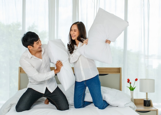 Jovem casal asiático atraente feliz provocando um ao outro com uma luta de almofadas na cama.