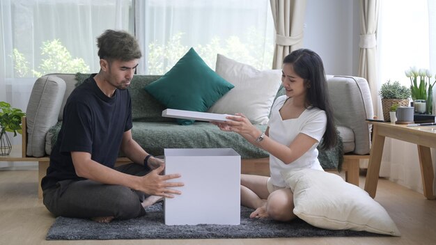 Jovem casal asiático sentado no chão e desembalando caixas de papelão na nova casa Conceito de remoções de renovação de casas de realocação