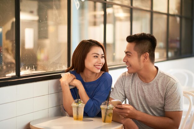 Jovem casal asiático sorridente, sentado com smartphones à mesa pequena e conversando em um café moderno