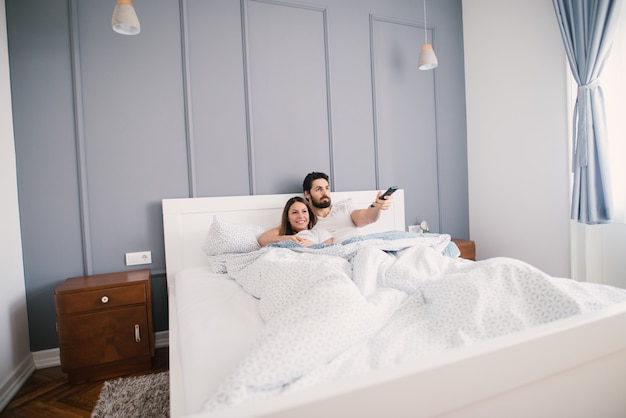 Jovem casal assistindo TV enquanto estava deitado na cama se abraçou.