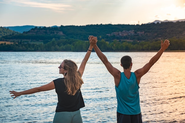 Jovem casal atraente abre os braços de mãos dadas em um belo lago no verão durante o pôr do sol descobrir