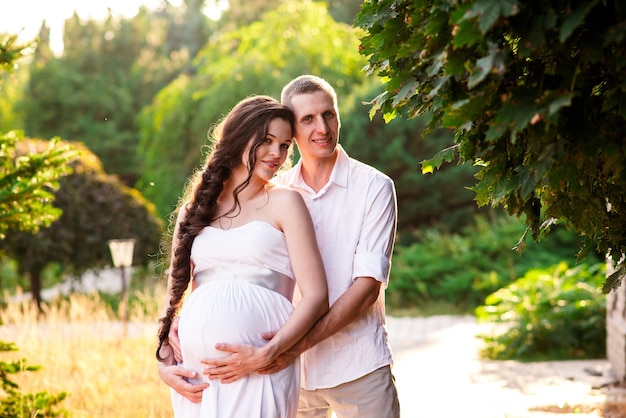 Jovem casal branco grávido romântico feliz no vestido de camisa branca beijando abraçando a barriga tocando na natureza no parque de pinheiros Mulher grávida esperando um bebê Família futura mãe e pai