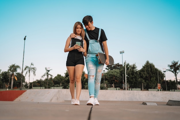 Jovem casal caminhando, carregando um skate e olhando para o celular no parque.