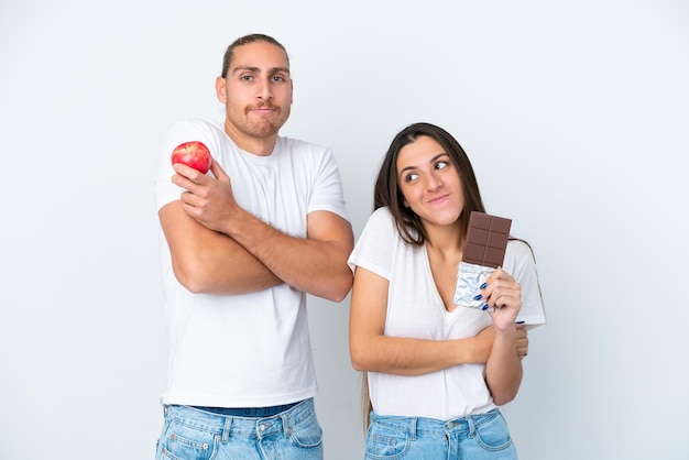 Jovem casal caucasiano com chocolate e maçã isolado no fundo branco fazendo gesto de dúvidas enquanto levanta os ombros