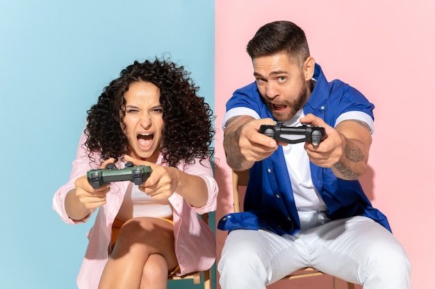 Foto jovem casal caucasiano emocional em roupas casuais brilhantes, posando em fundo rosa e azul. conceito de emoções humanas, expressão facial, relações, anúncio. homem e mulher jogam videogame, tentam vencer.