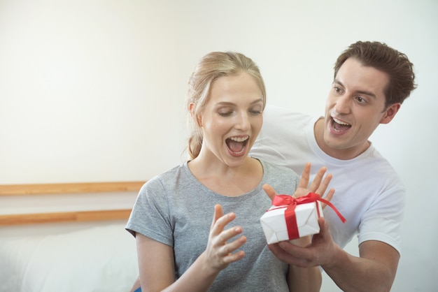 Jovem casal caucasiano feliz comemorando em casa. homem bonito namorado dá sua namorada com caixa de presente surpreendentemente.