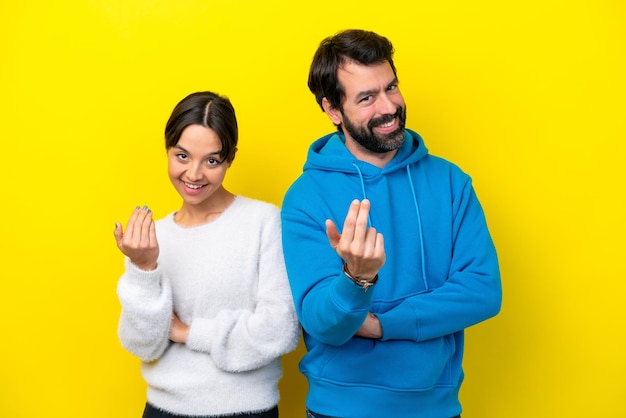 Jovem casal caucasiano isolado em fundo amarelo convidando para vir com a mão