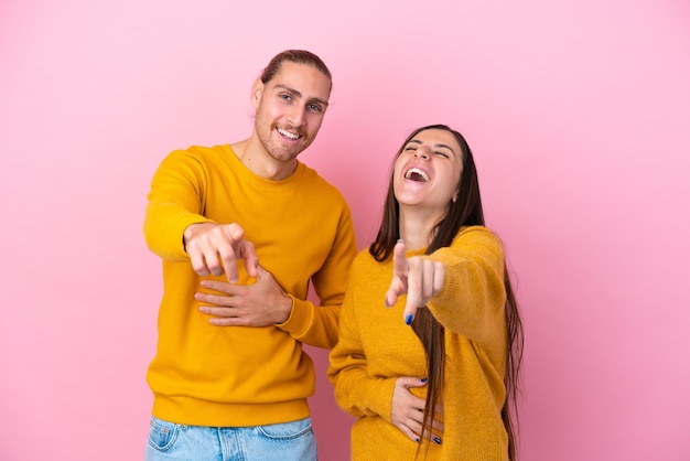 Jovem casal caucasiano isolado em fundo rosa apontando com o dedo para alguém e rindo muito