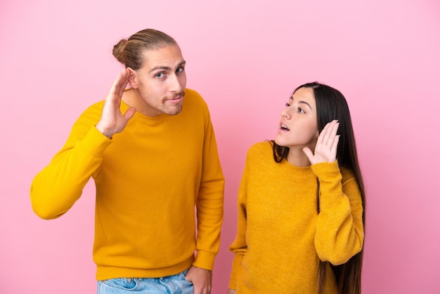 Jovem casal caucasiano isolado em fundo rosa ouvindo algo colocando a mão na orelha