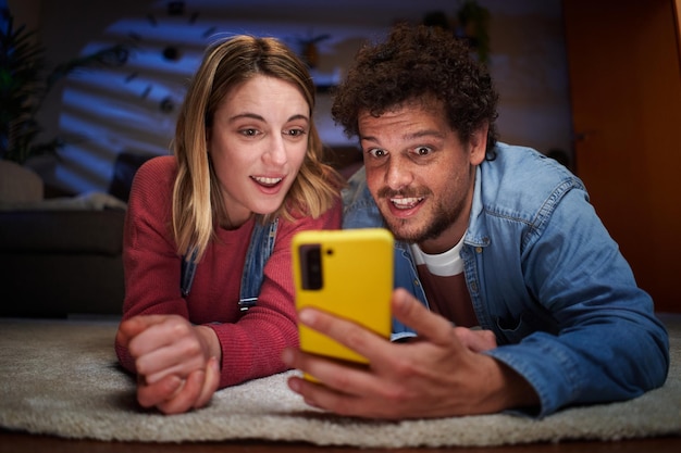 Jovem casal caucasiano olhando com rosto surpreso para o telefone deitado no tapete na sala de estar à noite