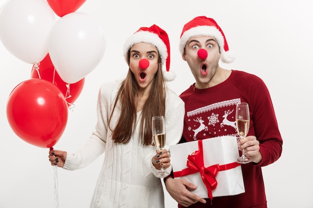 Jovem casal caucasiano segurando presentes, champnage e balão fazendo uma cara engraçada no natal.