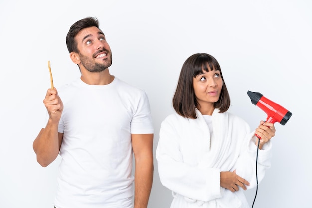 Jovem casal caucasiano segurando um secador de cabelo e escova de dentes isolado no fundo branco olhando para cima enquanto sorria