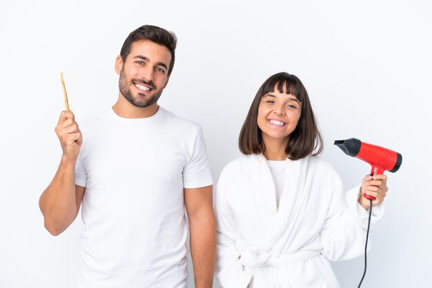 Foto jovem casal caucasiano segurando um secador de cabelo e escova de dentes isolado no fundo branco posando com os braços no quadril e sorrindo