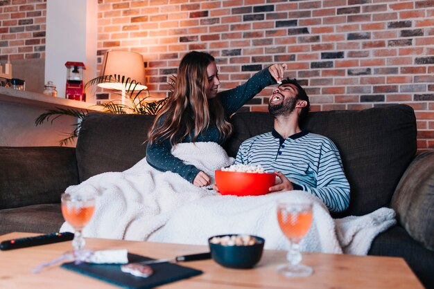 Foto jovem casal caucasiano sentado em um sofá cinza enquanto brincava com pipoca na boca