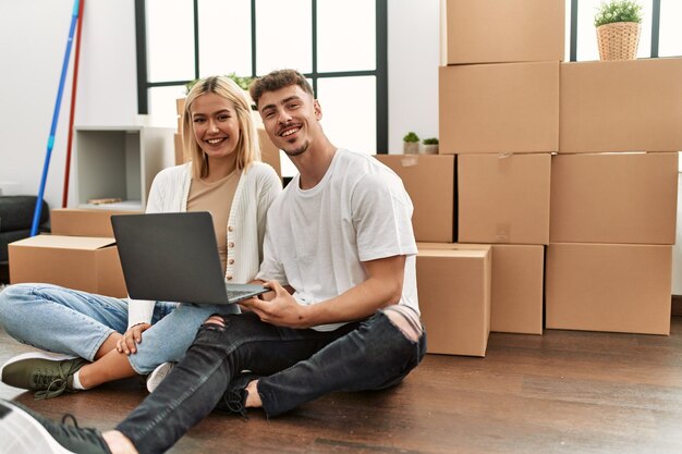 Jovem casal caucasiano sorrindo feliz usando laptop sentado na nova casa.