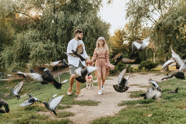 Jovem casal com cachorro está se divertindo e brincando espalhando pombos no parque primavera