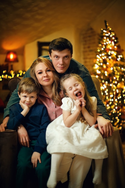 Jovem casal com dois filhos posando em uma cadeira em um interior de natal com uma árvore de natal