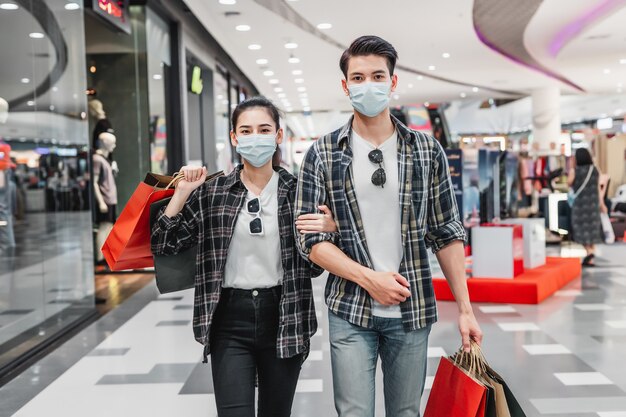 Jovem casal com máscara de proteção segurando várias sacolas de papel andando no corredor de um grande shopping center