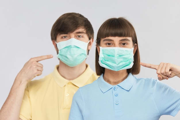 Foto jovem casal com máscara facial protetora olhando para a câmera e apontando para ela, proteção anti-coronavírus