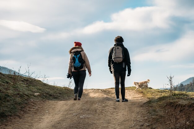 Jovem casal com mochilas e um cachorro andando nas montanhas, homem e mulher calorosamente vestidos de caminhada.