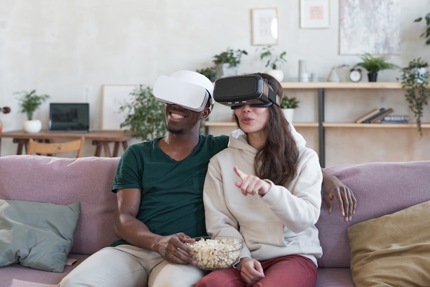 Jovem casal com óculos de RV sentado no sofá com pipoca e assistindo filme em realidade virtual