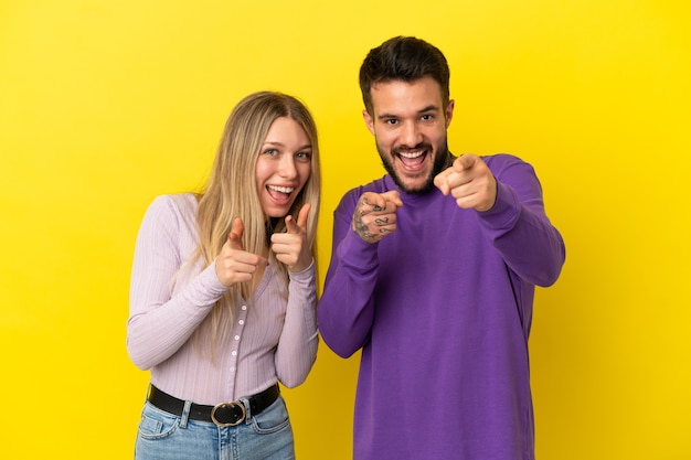 Jovem casal com um fundo amarelo isolado apontando o dedo para você