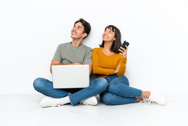 Jovem casal com um laptop e celular sentado no chão, olhando para cima enquanto sorri