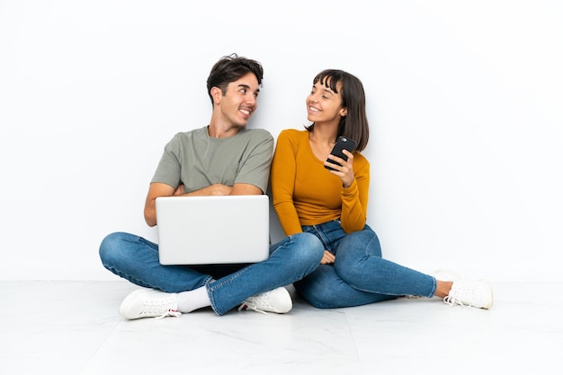 Jovem casal com um laptop e celular sentado no chão, olhando por cima do ombro com um sorriso