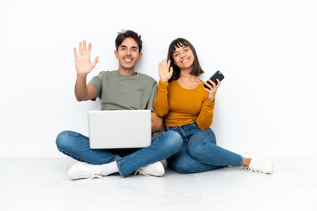 Jovem casal com um laptop e celular sentado no chão saudando com a mão com expressão feliz