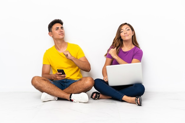 Jovem casal com um laptop sentado no chão branco orgulhoso e satisfeito