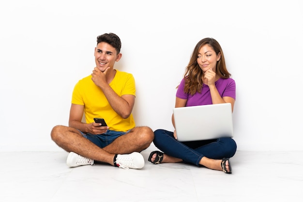 Jovem casal com um laptop sentado no chão, olhando para o lado e sorrindo