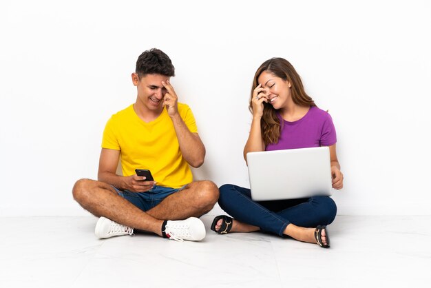 Jovem casal com um laptop sentado no chão, rindo branco