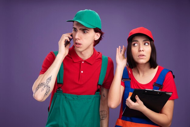 Jovem casal com uniforme de trabalhador da construção civil e boné preocupado falando no telefone olhando para baixo uma garota curiosa segurando o lápis e a prancheta, olhando para o lado, ouvindo uma conversa telefônica isolada