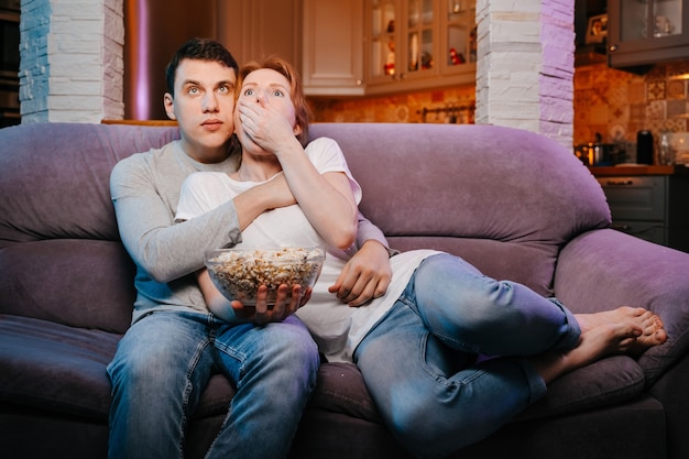 Jovem casal comendo pipoca e assistindo a um filme em casa no sofá muito assustado