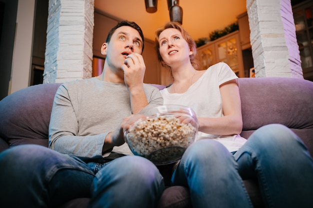Jovem casal comendo pipoca e assistindo a um filme em casa no sofá muito assustado