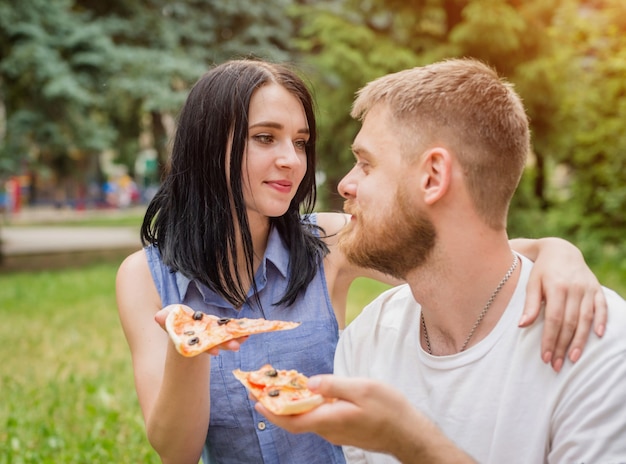 Jovem casal comendo pizza no parque. Eles se tratam e riem.