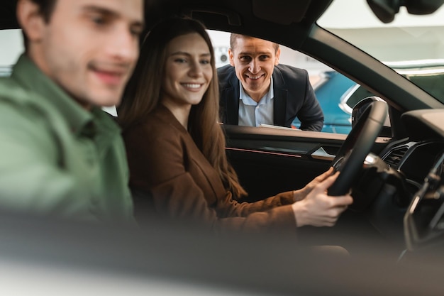 Jovem casal comprando carro novo sentado no salão de automóveis fazendo test drive conversando com vendedor na concessionária de automóveis