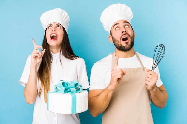 Jovem casal cozinhando um bolo isolado apontando para cima com a boca aberta.