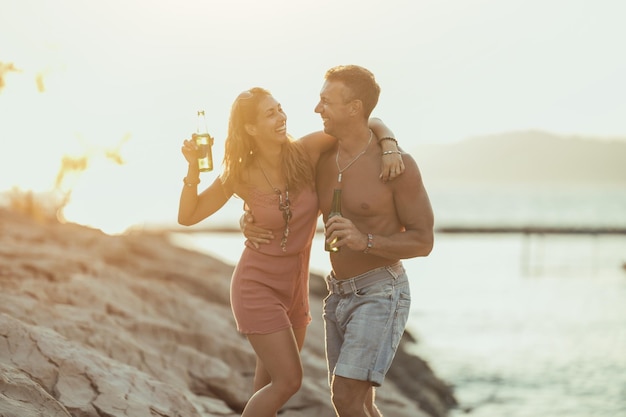 Jovem casal dançando e bebendo cerveja na praia à beira-mar.