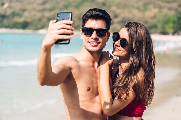 Jovem casal de amantes românticos relaxando juntos na praia tropical. Homem e mulher tirando uma selfie com smartphone e curtindo a vida. Férias de verão