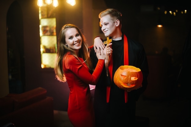 Jovem casal de estudantes universitários vestidos de padre e bruxa em uma festa de Halloween. Jovem casal posando com uma abóbora nas mãos e olhando para a câmera
